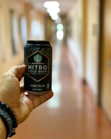 A hand holding a can of Starbucks Nitro Cold Brew is prominently featured, with a blurred hallway in the background. The design on the can is detailed, and the background is softly out of focus, indicating an indoor setting.