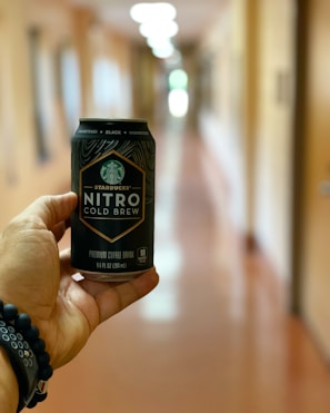 A hand holding a can of Starbucks Nitro Cold Brew is prominently featured, with a blurred hallway in the background. The design on the can is detailed, and the background is softly out of focus, indicating an indoor setting.