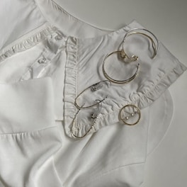 An arrangement of several pieces of metal jewelry, including hoop earrings and a ring, laid out on a white collared shirt with button and ruffle details.