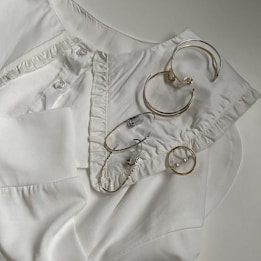 An arrangement of several pieces of metal jewelry, including hoop earrings and a ring, laid out on a white collared shirt with button and ruffle details.