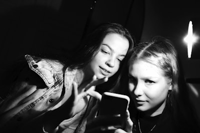 Two young women are looking at a smartphone together. One is holding the phone while the other is pointing at the screen. The scene is lit by a harsh light source creating high contrast and casting deep shadows.