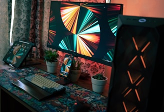 A tidy and modern computer setup includes a large computer monitor displaying vibrant abstract art, a keyboard with colorful keycaps, and a mouse on a graphic-patterned mouse pad. There is also a tablet and a smartphone displaying the same abstract art. The workspace includes small potted plants and is illuminated by warm lighting, with decorative curtains in the background.