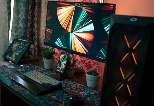A tidy and modern computer setup includes a large computer monitor displaying vibrant abstract art, a keyboard with colorful keycaps, and a mouse on a graphic-patterned mouse pad. There is also a tablet and a smartphone displaying the same abstract art. The workspace includes small potted plants and is illuminated by warm lighting, with decorative curtains in the background.
