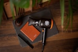 A wooden surface displays a hairdryer, a leather notebook with the words 'Live to Create', a black comb, and a small wooden container. A slate or dark mat serves as a background for these items, with green plant leaves partially visible on the right side.