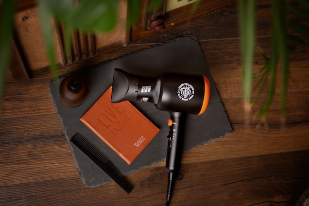 A wooden surface displays a hairdryer, a leather notebook with the words 'Live to Create', a black comb, and a small wooden container. A slate or dark mat serves as a background for these items, with green plant leaves partially visible on the right side.