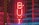 A bright red neon sign reading 'BUY' is centered in the image, placed in front of a backdrop featuring a partially visible American flag with stars and stripes. The overall scene has a dark, moody ambiance accentuated by the glowing neon light.