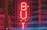 A bright red neon sign reading 'BUY' is centered in the image, placed in front of a backdrop featuring a partially visible American flag with stars and stripes. The overall scene has a dark, moody ambiance accentuated by the glowing neon light.