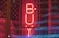 A bright red neon sign reading 'BUY' is centered in the image, placed in front of a backdrop featuring a partially visible American flag with stars and stripes. The overall scene has a dark, moody ambiance accentuated by the glowing neon light.