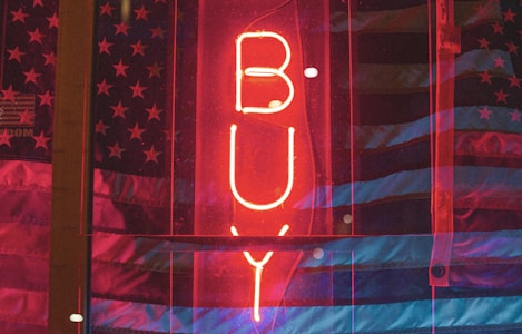A bright red neon sign reading 'BUY' is centered in the image, placed in front of a backdrop featuring a partially visible American flag with stars and stripes. The overall scene has a dark, moody ambiance accentuated by the glowing neon light.