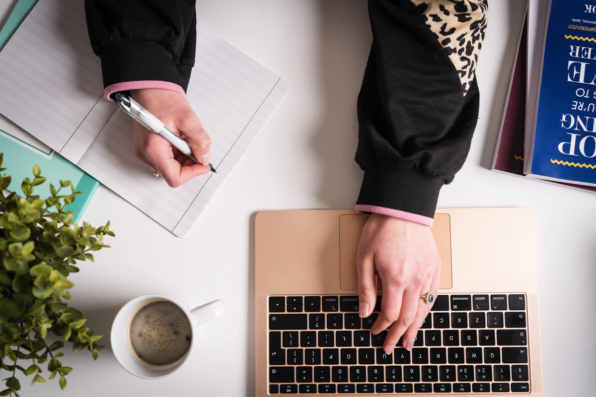 A desk from above: one hand writing in a notebook, the other typing on a laptop