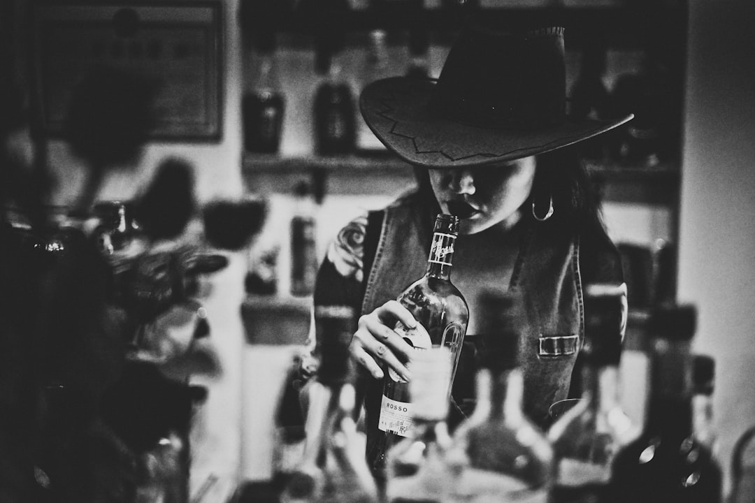 grayscale photo of man in black hat holding glass bottle