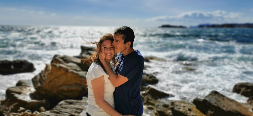 man in blue shirt hugging woman in white shirt near sea during daytime