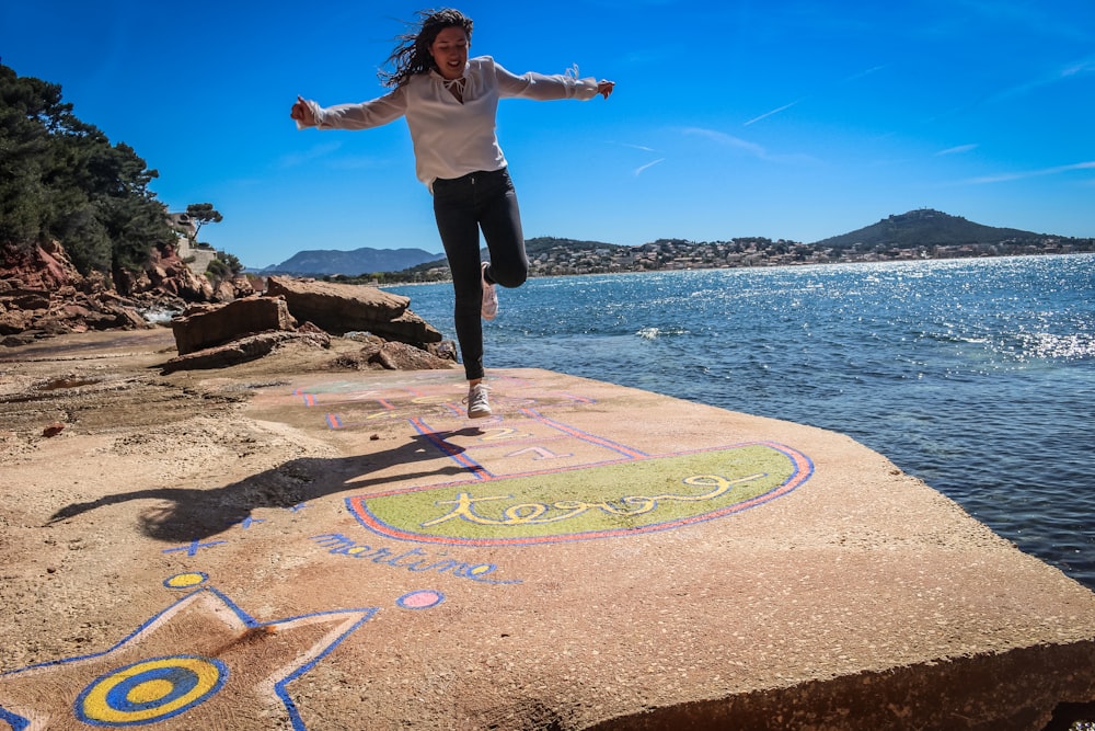 woman in white shirt and black pants standing on rock near body of water during daytime