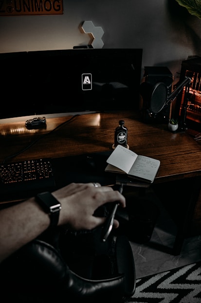 A dark and moody workspace with a sleek wooden desk featuring a computer setup with two large monitors. A microphone is positioned to the right of the desk, and a bottle labeled 'Black Pepper Coffee' is placed next to an open notebook with handwritten notes. A hand wearing a smartwatch is in the foreground, appearing to reach for something. There are some geometric hexagonal wall decorations and a 'UNIMOG' sign partially visible on the wall.