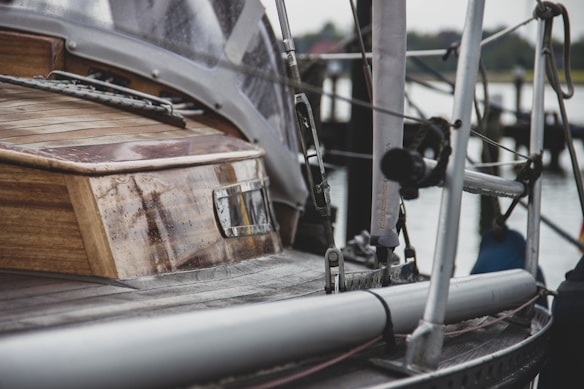 A close-up view of a sailboat's deck, highlighting the wooden cabin structure and metal rigging. The image captures the texture of the wood and metal, with a slightly blurred background of a marina and water.