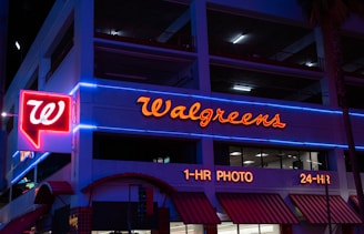 The image displays the exterior of a Walgreens store at night, featuring prominently illuminated signage and red neon lights accompanied by blue neon trim on the building. The storefront includes signs indicating services such as 1-hour photo and 24-hour availability.