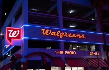 The image displays the exterior of a Walgreens store at night, featuring prominently illuminated signage and red neon lights accompanied by blue neon trim on the building. The storefront includes signs indicating services such as 1-hour photo and 24-hour availability.