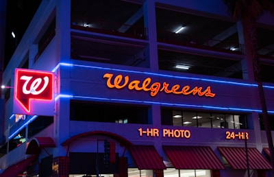 The image displays the exterior of a Walgreens store at night, featuring prominently illuminated signage and red neon lights accompanied by blue neon trim on the building. The storefront includes signs indicating services such as 1-hour photo and 24-hour availability.
