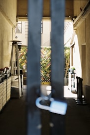 A gated passageway with large vertical metal bars in the foreground and a lock hanging on one of them. Beyond the gate, there is a partially visible outdoor space with potted plants and a heater. The walls on either side are beige, and there is an intricate wooden fence adorned with greenery at the end of the passage.