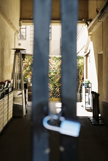 A gated passageway with large vertical metal bars in the foreground and a lock hanging on one of them. Beyond the gate, there is a partially visible outdoor space with potted plants and a heater. The walls on either side are beige, and there is an intricate wooden fence adorned with greenery at the end of the passage.