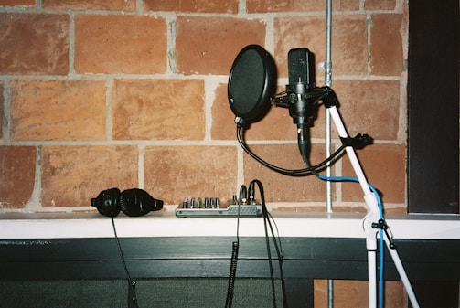 A recording studio setup featuring a microphone mounted on a stand with a pop filter, a pair of headphones, and an audio mixer resting on a white shelf. The background is a brick wall, giving the scene a rustic and professional studio vibe.
