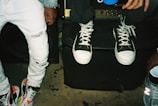 A close-up of two individuals wearing casual clothing. One person is seated on a speaker wearing white sneakers with black accents and ripped jeans. The other person stands next to them, wearing multicolored sneakers and lighter jeans. The setting appears to be casual and informal.