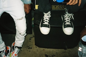 A close-up of two individuals wearing casual clothing. One person is seated on a speaker wearing white sneakers with black accents and ripped jeans. The other person stands next to them, wearing multicolored sneakers and lighter jeans. The setting appears to be casual and informal.