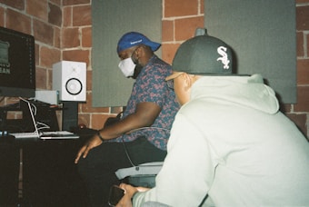 Two individuals are seated in a recording studio. One person, wearing a blue baseball cap and floral shirt, is focusing on a computer screen. The other person, wearing a gray hoodie and black baseball cap, is nearby holding a smartphone. The room features exposed brick walls, soundproof panels, and studio equipment such as a speaker and computer monitor.