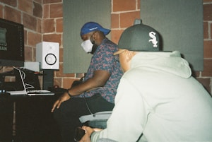 Two individuals are seated in a recording studio. One person, wearing a blue baseball cap and floral shirt, is focusing on a computer screen. The other person, wearing a gray hoodie and black baseball cap, is nearby holding a smartphone. The room features exposed brick walls, soundproof panels, and studio equipment such as a speaker and computer monitor.
