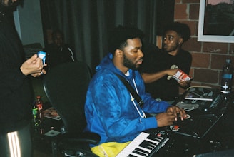 A recording studio environment with three people. One person is sitting in front of a keyboard and computer equipment, wearing a blue hoodie and yellow pants, seemingly focused on the task at hand. Another person, wearing a black shirt, is holding a bag of snacks, looking towards the person in the hoodie. A third person, partially visible, is holding a smartphone in their hand. Various items, including a water bottle and snacks, are visible on the table.