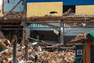 The image depicts a partially demolished building with debris scattered around. The upper part of the building has yellow and blue painted walls, with significant damage and rubble. Broken signs, bent metal, and various fragments of construction materials are visible. A no-parking sign is seen in the foreground.