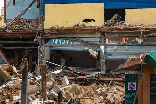 The image depicts a partially demolished building with debris scattered around. The upper part of the building has yellow and blue painted walls, with significant damage and rubble. Broken signs, bent metal, and various fragments of construction materials are visible. A no-parking sign is seen in the foreground.