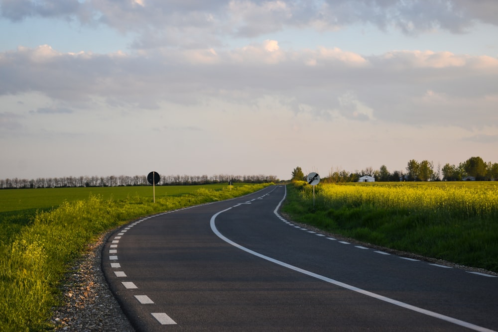 gray asphalt road between green grass field under white cloudy sky during daytime