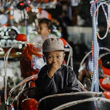 A young child wearing a hat sits in a small, toy vehicle surrounded by colorful lights. The child appears thoughtful, with their hand near their mouth. In the background, another child is visible, also in a toy vehicle.