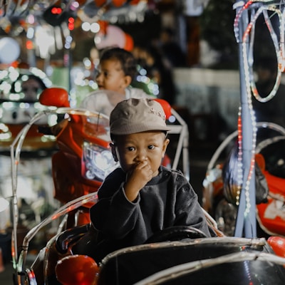 A young child wearing a hat sits in a small, toy vehicle surrounded by colorful lights. The child appears thoughtful, with their hand near their mouth. In the background, another child is visible, also in a toy vehicle.