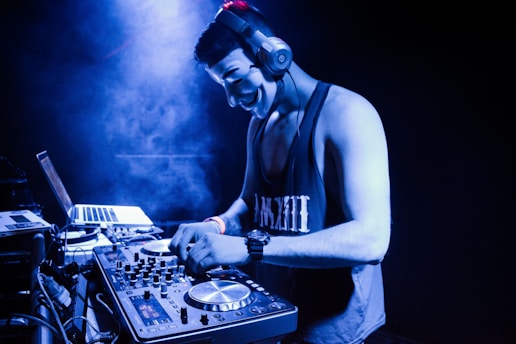 A DJ wearing headphones and a mask is concentrating on mixing music. They stand behind a DJ controller with various knobs and dials, beside a laptop. The setting is dimly lit with blue lighting and a slight haze, suggesting an energetic or mysterious atmosphere.