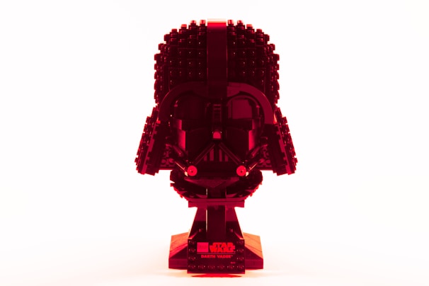 A Lego model resembling the helmet of a dark character with a glossy finish, presented in a striking red hue against a bright white background. The Lego pieces are intricately assembled to capture the iconic shape and features of the helmet, with a base displaying related branding.
