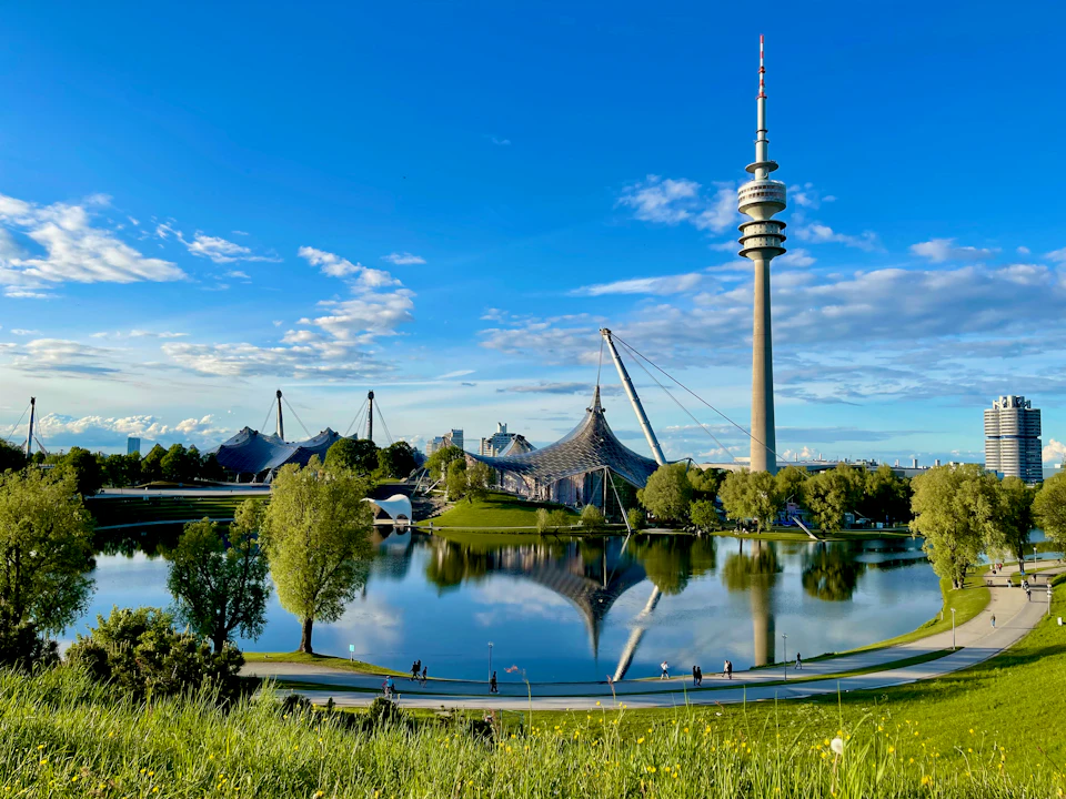 The image depicts a large, circular lake surrounded by lush green trees and grassy areas. The calm waters of the lake reflect the clear blue sky above, creating a serene atmosphere. In the background, there is a tall tower with a pointed top that stands out against the sky. On the left side of the image, several tents are visible, suggesting an event or gathering taking place nearby.