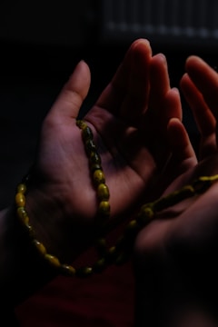 Hands are gently cupped together, holding a string of prayer beads. The lighting is dim, casting soft shadows, and highlighting the texture of the skin and beads.