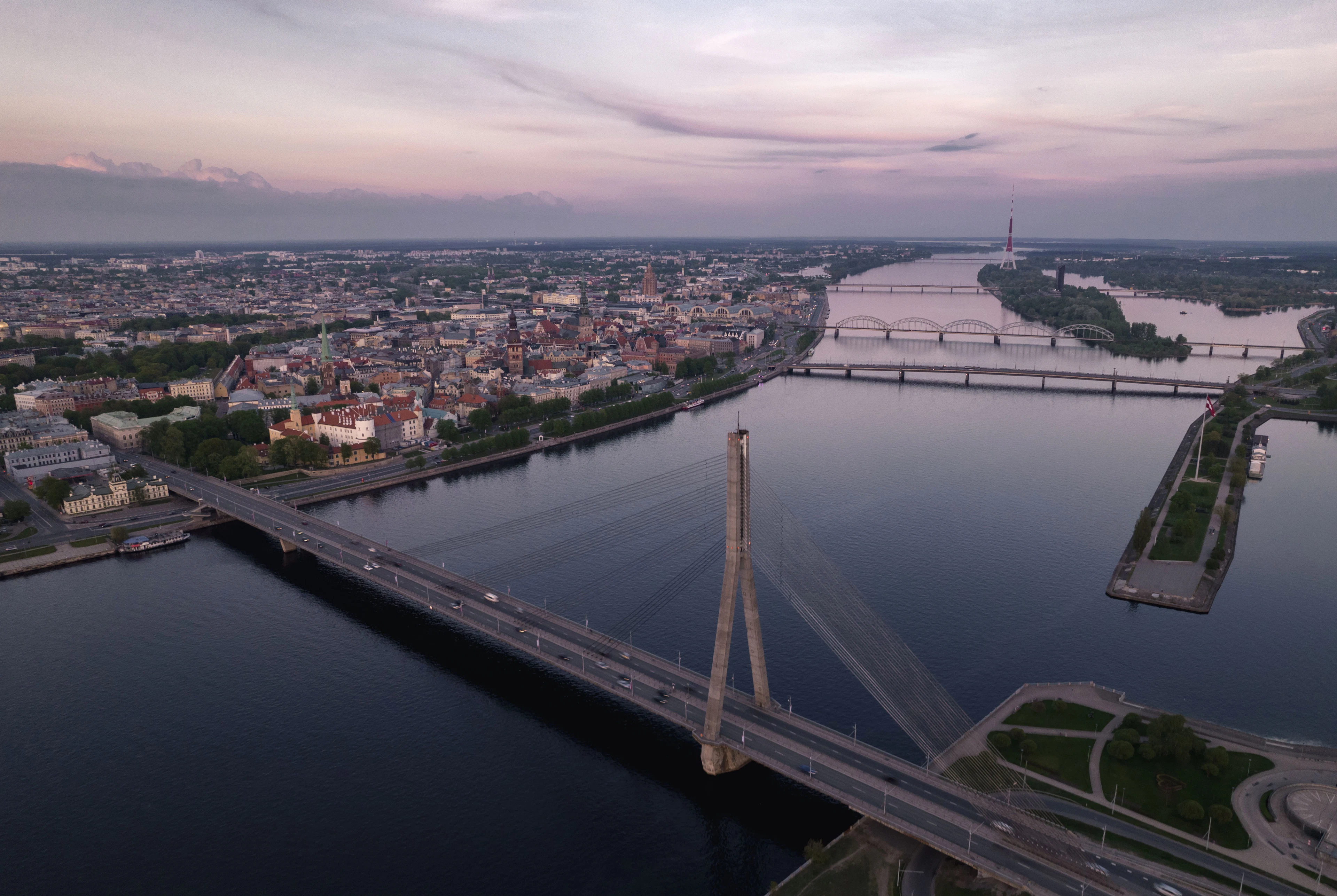 The cable-stayed Vansu Bridge sweeping across the Daugava at dusk, with Riga's old town spread along the far bank and the river's other bridges receding under a pink and lilac sky.