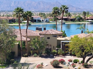 A luxurious residential area featuring a villa with a tile roof surrounded by palm trees and lush landscaping. In the background, a tranquil lake reflects the clear blue sky, and mountains rise up in the distance, creating a serene and picturesque setting.