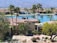 A luxurious residential area featuring a villa with a tile roof surrounded by palm trees and lush landscaping. In the background, a tranquil lake reflects the clear blue sky, and mountains rise up in the distance, creating a serene and picturesque setting.