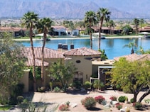 A luxurious residential area featuring a villa with a tile roof surrounded by palm trees and lush landscaping. In the background, a tranquil lake reflects the clear blue sky, and mountains rise up in the distance, creating a serene and picturesque setting.