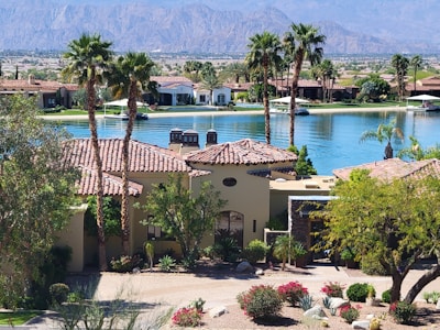A luxurious residential area featuring a villa with a tile roof surrounded by palm trees and lush landscaping. In the background, a tranquil lake reflects the clear blue sky, and mountains rise up in the distance, creating a serene and picturesque setting.