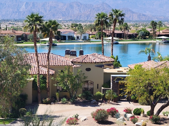 A luxurious residential area featuring a villa with a tile roof surrounded by palm trees and lush landscaping. In the background, a tranquil lake reflects the clear blue sky, and mountains rise up in the distance, creating a serene and picturesque setting.