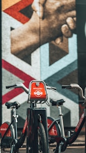 A row of Santander rental bicycles is parked in front of a large mural featuring an image of two hands gripping each other firmly. The mural consists of geometric patterns with various shades of color, adding a vibrant background to the scene.
