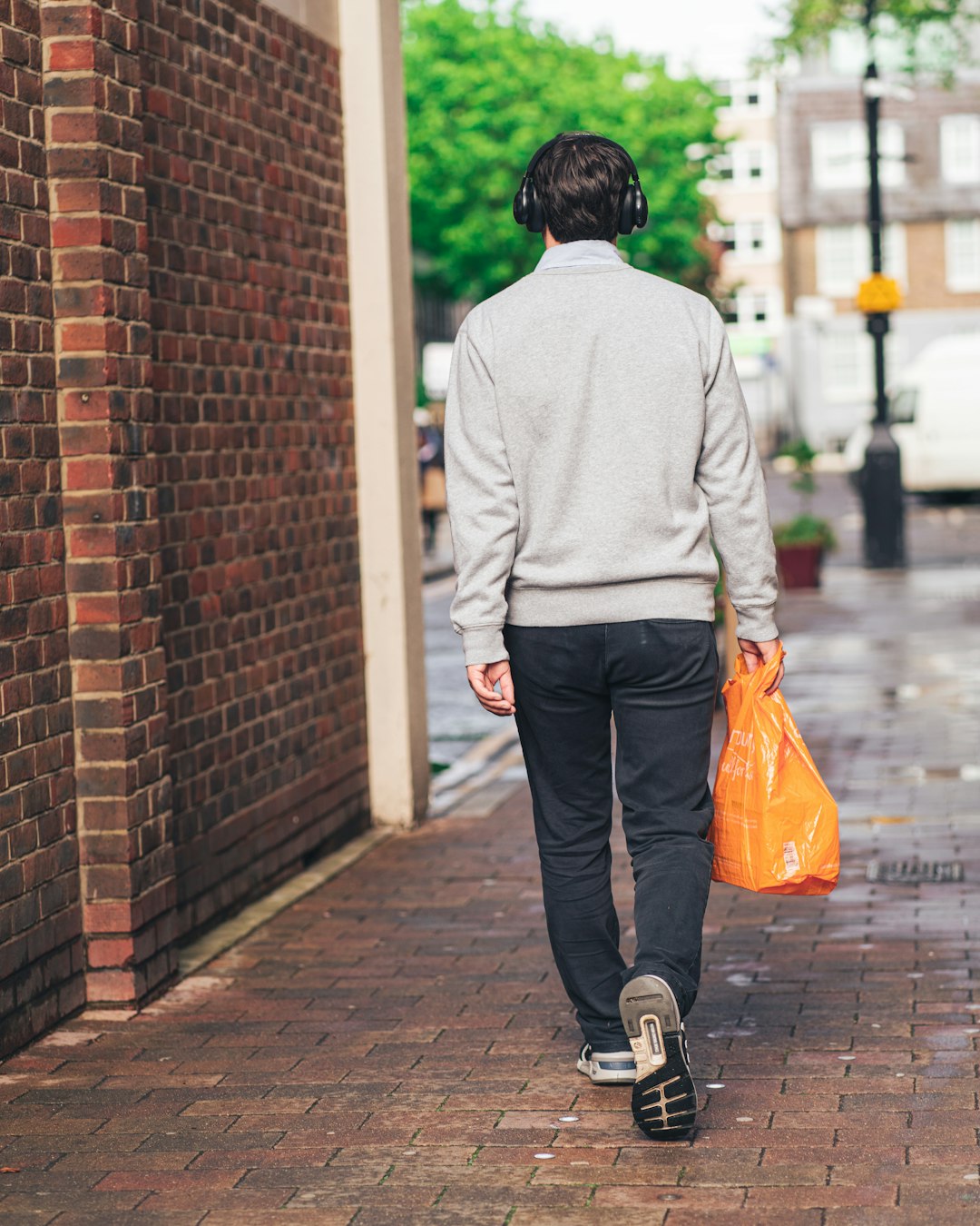 man in gray hoodie and black pants holding orange plastic bag walking on sidewalk during daytime