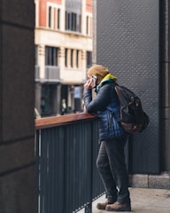 A person stands on a city balcony, leaning against a railing while talking on a mobile phone. They are dressed in a blue jacket and brown beanie, wearing a backpack. The background features urban architecture with tall buildings.