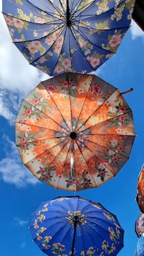Colorful umbrellas with floral patterns are seen from below against a bright blue sky. The umbrellas are open and showcase intricate designs, primarily featuring flowers and leaves, with colors such as blue, orange, and green.