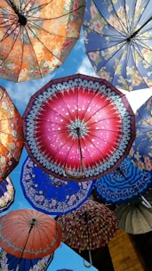 A collection of colorful umbrellas with intricate patterns and designs is opened against a backdrop of blue sky. The umbrellas vary in color, including shades of red, blue, orange, and purple, and feature floral and geometric motifs.
